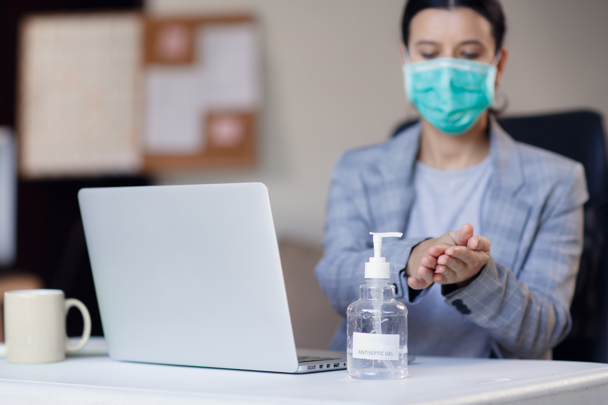 Woman wearing face mask while working in office with laptop and using hand alcohol gel or sanitizer bottle dispenser against corona virus Covid-19. Antiseptic, hygiene and health concept. Prevention of the pandemic virus outbreak