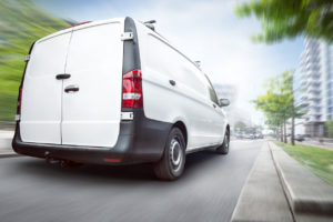 Rear view of a white commercial van driving in a city. Motion blurred city background with buildings and trees.