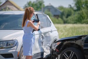 A stressed female driver captures photos of a vehicle collision on her mobile phone at the roadside after an accident, emphasizing road safety and insurance claims. 