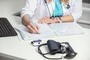 A close-up of a general practitioner’s hands filling out medical records. The doctor, in a white coat, writes notes, prepares documents, and works with a laptop, focusing on paperwork, reports, and prescriptions. 
