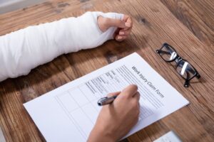 Close-up of a businesswoman with a bandaged hand completing a work injury claim form at a wooden desk.