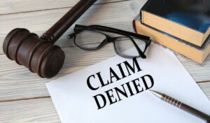 "CLAIM DENIED" written on white paper, set against a background featuring a judge's gavel, glasses, and a pen.
