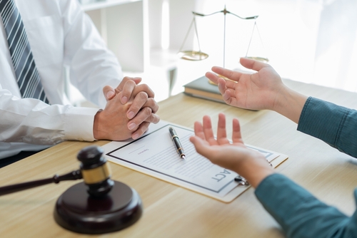 Two individuals in a legal consultation at a desk with a contract, gavel, and justice scale in view, discussing legal matters.
