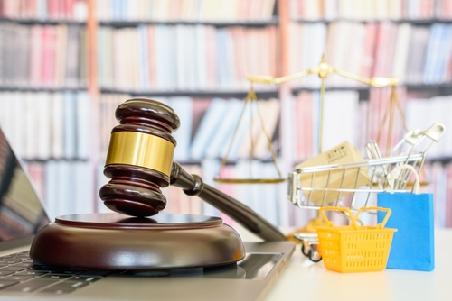A judge's gavel resting on a laptop next to a miniature shopping cart, scale of justice, and small shopping bags, symbolizing product liability law and consumer rights.