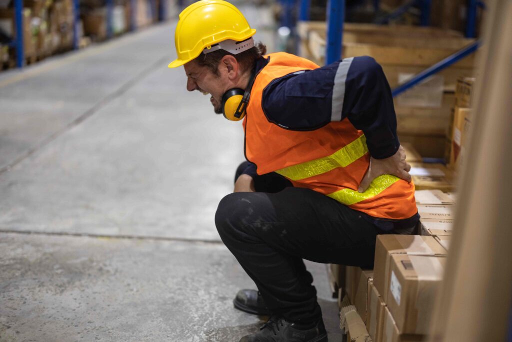 Warehouse worker wearing a hard hat and high-visibility vest sitting on boxes and holding his lower back in pain, illustrating a workplace back injury or lifting accident. Warehouse worker wearing a hard hat and high-visibility vest sitting on boxes and holding his lower back in pain, illustrating a workplace back injury or lifting accident.