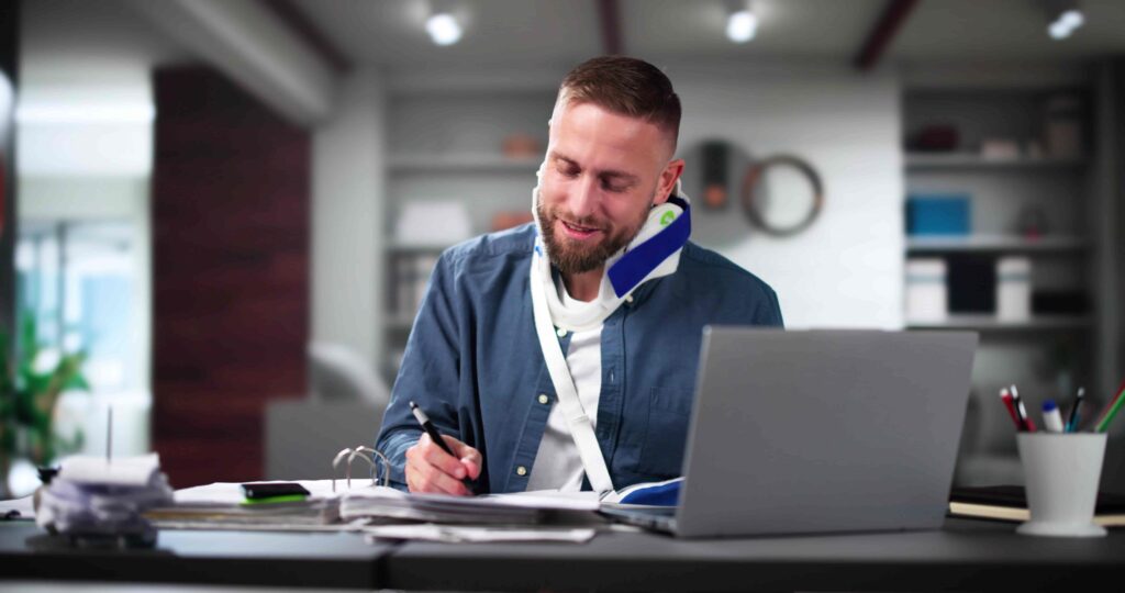 Injured office worker wearing a neck brace completing paperwork at a desk with a laptop, representing workplace injury recovery, workers’ compensation claim, and return-to-work process. Injured office worker wearing a neck brace completing paperwork at a desk with a laptop, representing workplace injury recovery, workers’ compensation claim, and return-to-work process.