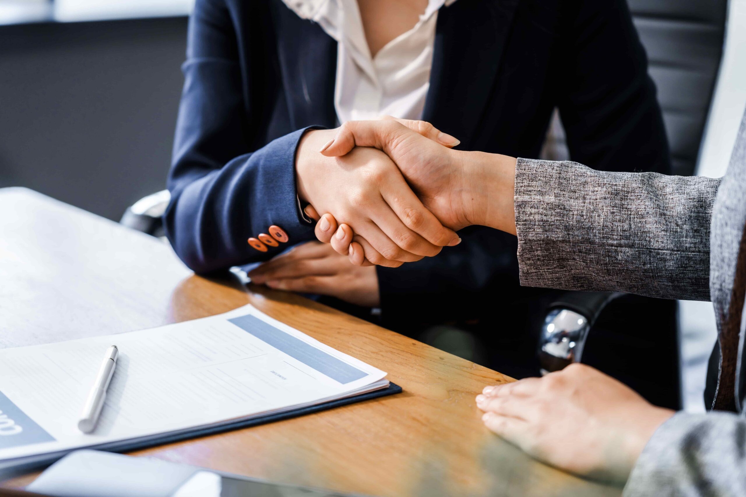 Business professionals shaking hands over a contract at a desk, symbolizing agreement, partnership, and successful business negotiation.