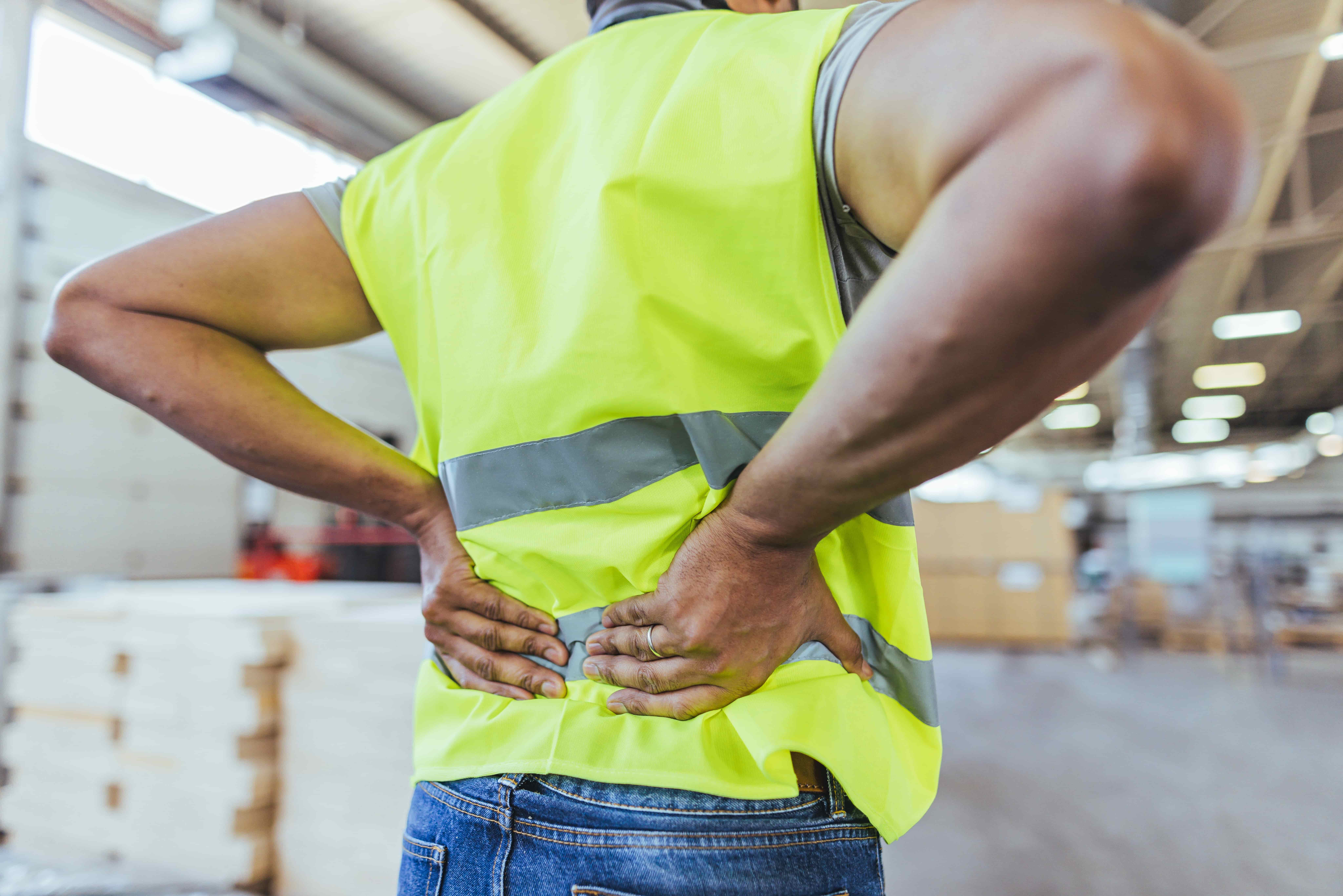 Warehouse worker wearing a high-visibility safety vest holding lower back in pain, representing workplace injury or back strain on the job.